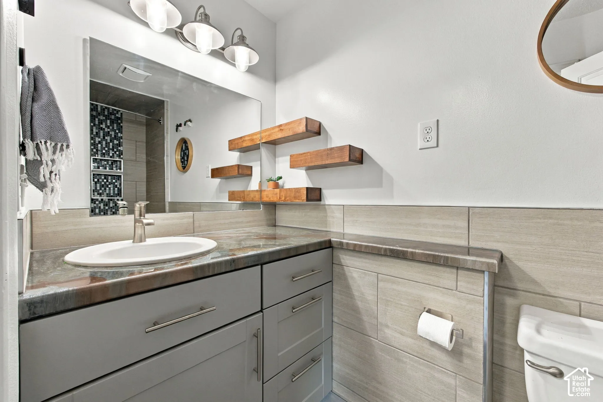Bathroom vanity featuring a grey cabinet, polished stone countertop with integrated sink, three-light brushed-metal fixture, wall-mounted wood-finish floating shelves, and tile-covered walls