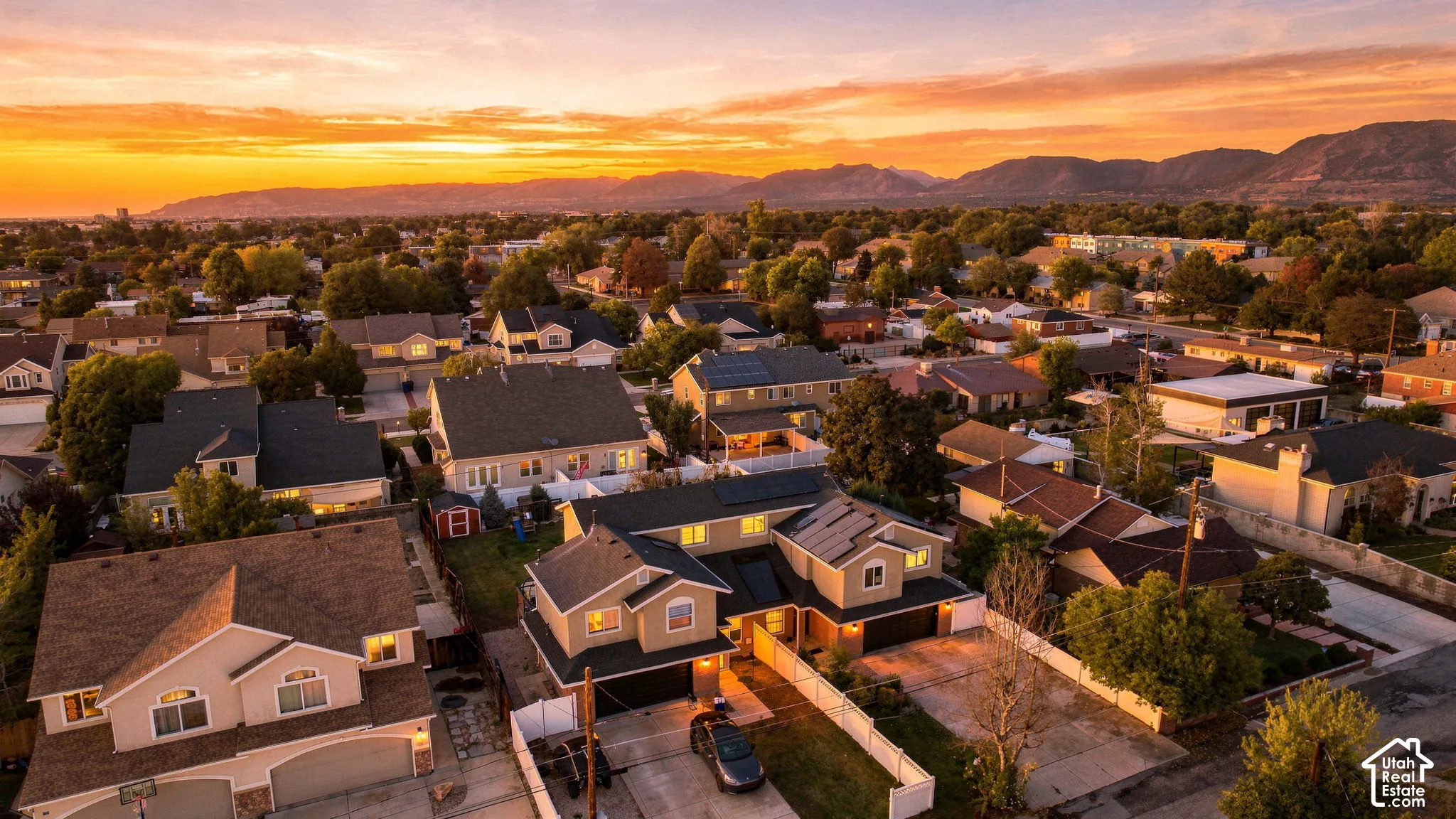 Aerial perspective featuring a multi-level residential structure with a shingled roof, roof-mounted solar panels, an attached multi-car garage, and a backdrop of mountain scenery at sunset