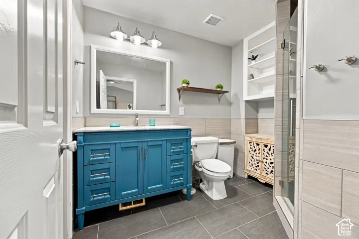 Blue vanity with hardware and stone countertop, large-format tile flooring, tile-surround shower enclosure, built-in shelving, and three-light vanity fixture