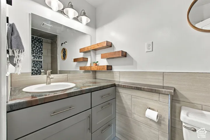 Bathroom vanity featuring a grey cabinet, polished stone countertop with integrated sink, three-light brushed-metal fixture, wall-mounted wood-finish floating shelves, and tile-covered walls