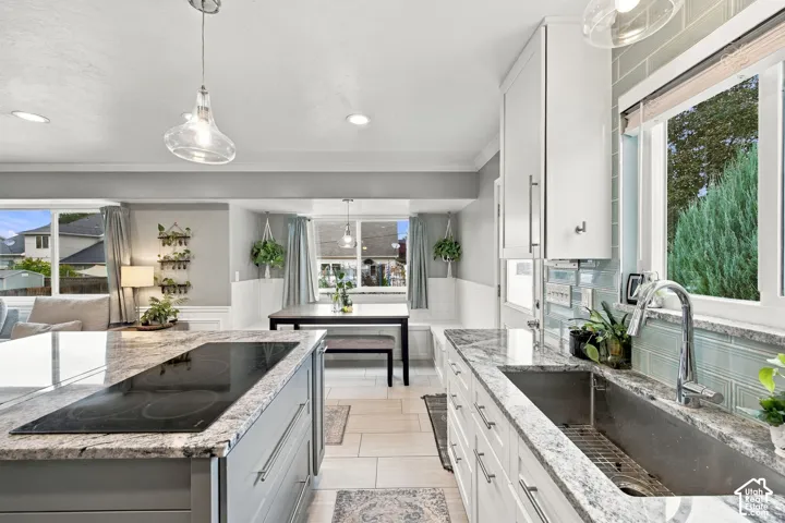 Kitchen featuring a large central island with an integrated cooktop, stone-finish countertops, stainless steel farmhouse-style sink, glass-tile backsplash, and glass pendant lighting