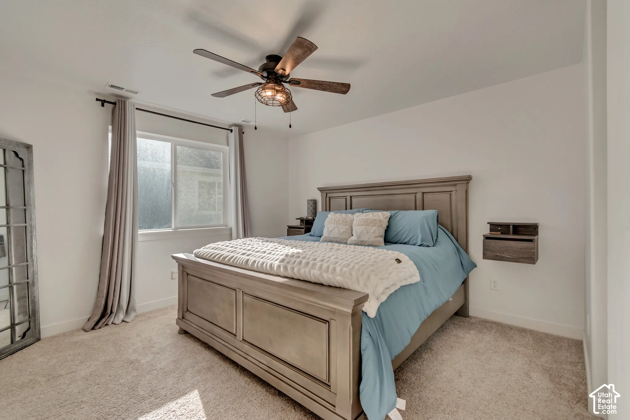 Carpeted flooring, white walls, window with roller shade, ceiling fan with integrated lighting, and a wall-mounted floating shelf