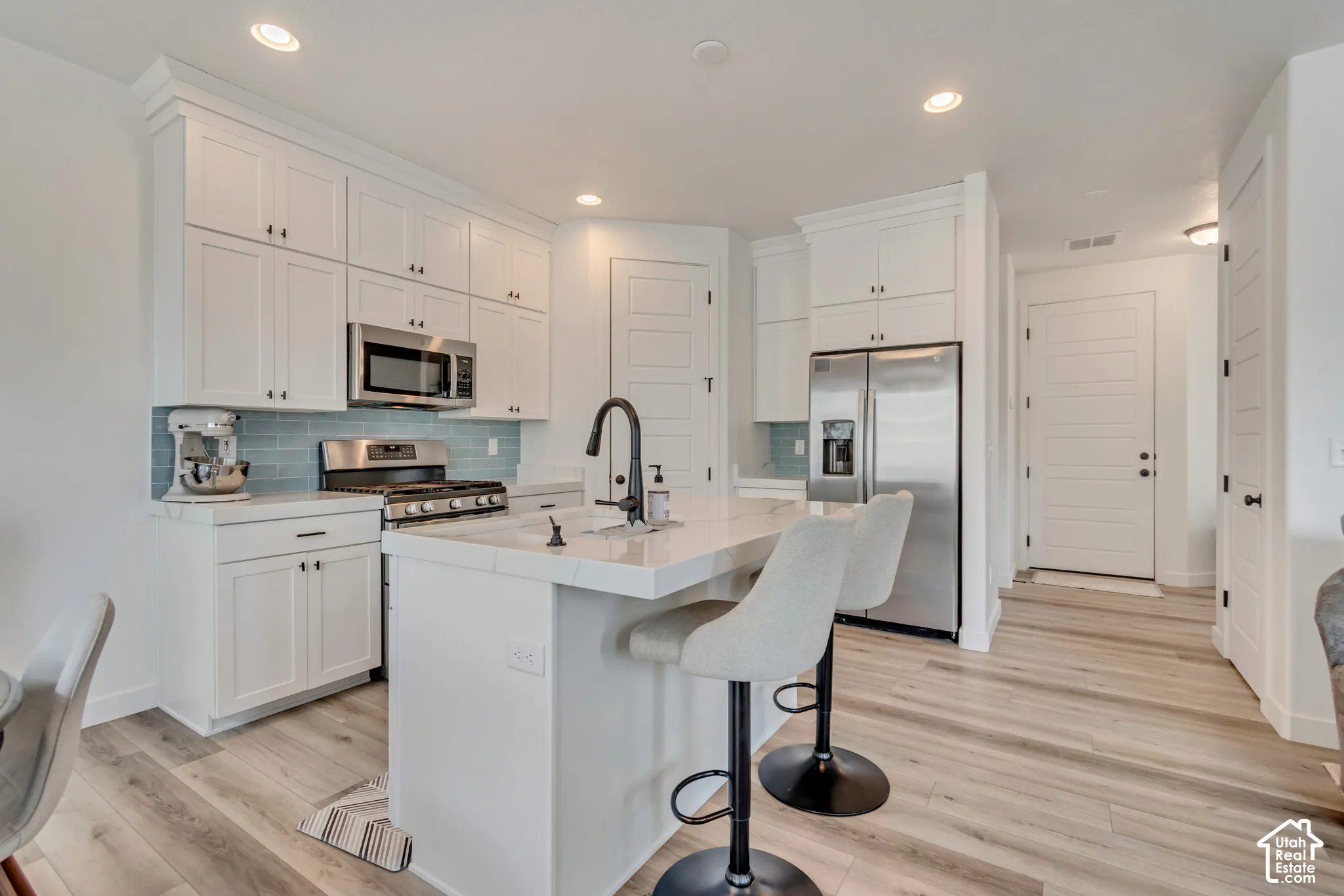 Bright kitchen featuring white shaker cabinetry, stainless steel appliances, light wood-finish flooring, a central island with countertop seating, and a blue subway tile backsplash