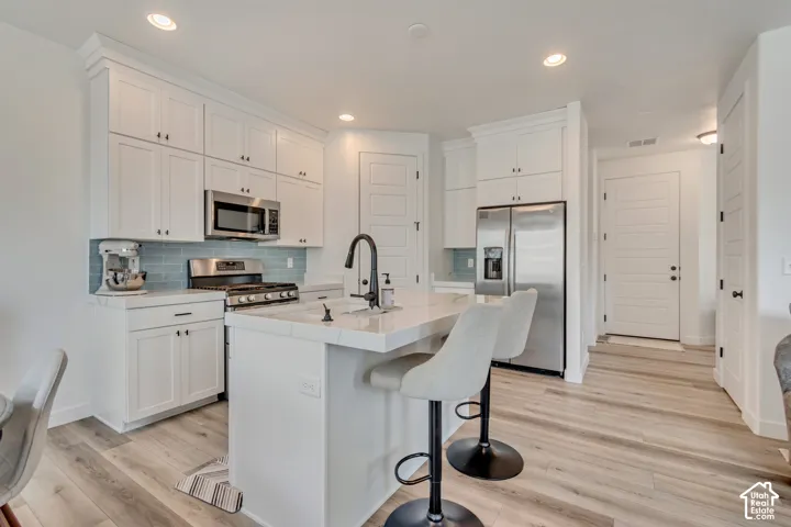Bright kitchen featuring white shaker cabinetry, stainless steel appliances, light wood-finish flooring, a central island with countertop seating, and a blue subway tile backsplash