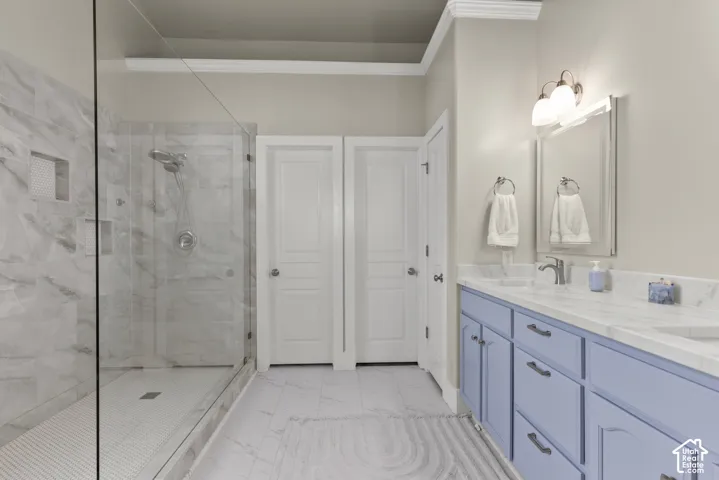 Walk-in glass-enclosed shower with stone-finish wall tile, dual-sink vanity featuring light-colored cabinetry, stone-finish countertop, crown molding, and framed wall mirror