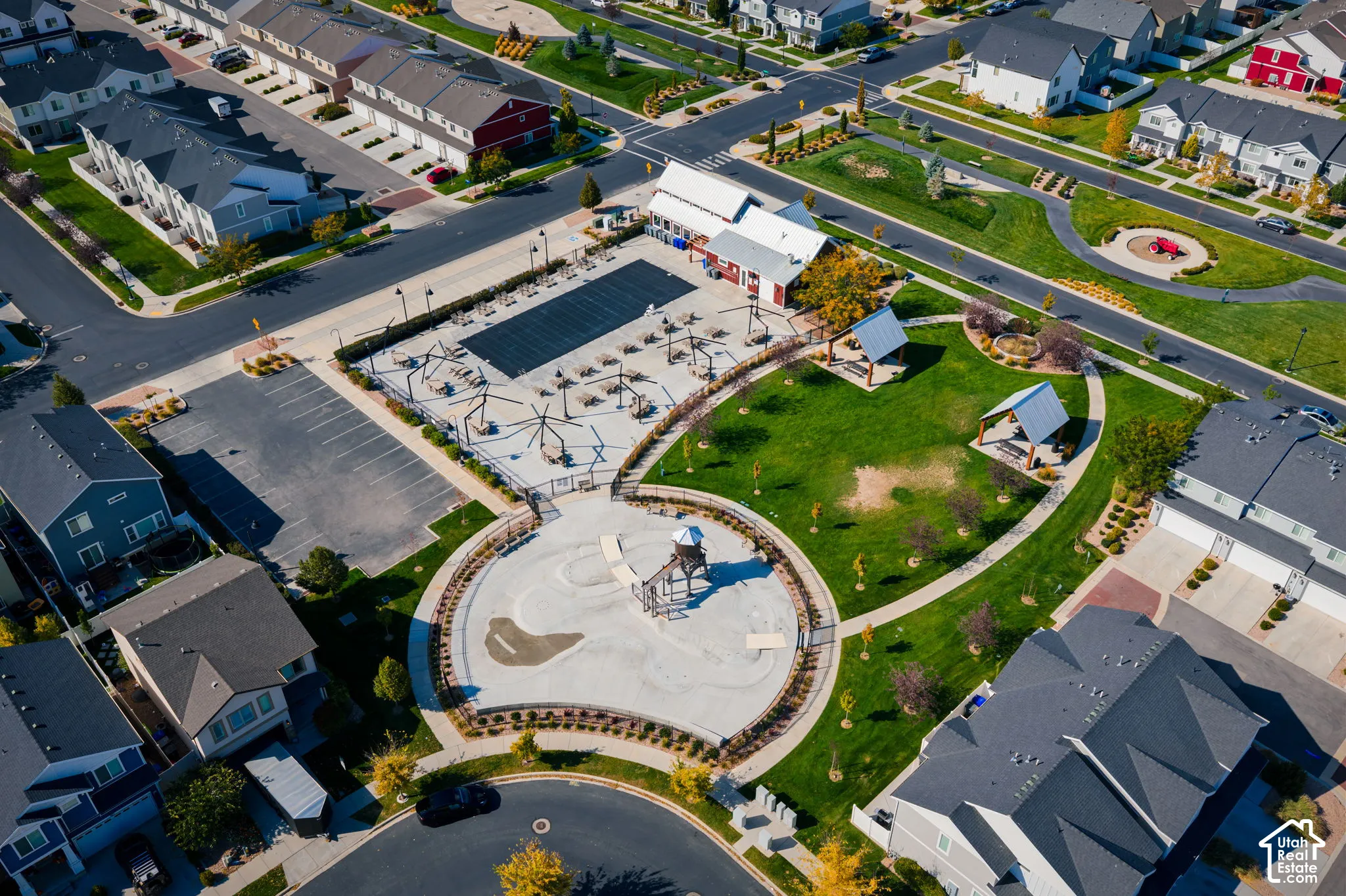 Aerial perspective of a community recreation area featuring a fenced swimming pool, paved parking lot, dedicated playground structure, open grass space, and covered pavilion seating