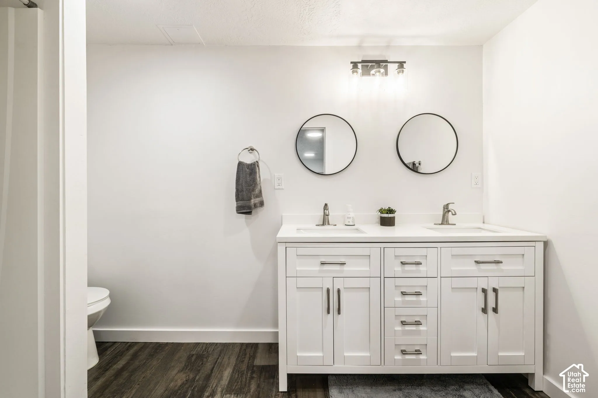 White dual-sink vanity with a solid surface countertop, metallic hardware, dual round mirrors, and a three-light fixture over wood-finish flooring