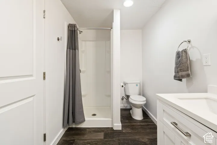 Bathroom featuring dark wood-finish flooring, white shaker-style vanity cabinet with an integrated sink, enclosed shower stall with built-in shelving, and white walls