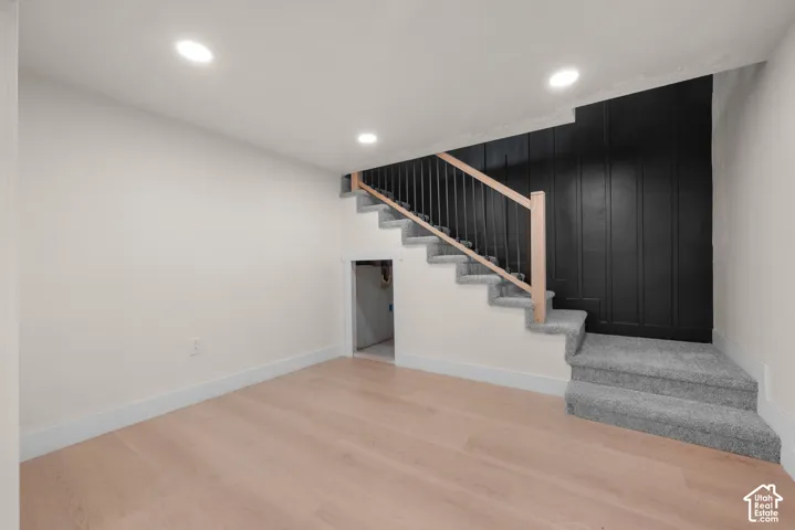 Staircase featuring carpeted treads, wood-finish handrails, black metal balusters, and a black paneled accent wall, complemented by recessed lighting and light-toned wood-finish flooring