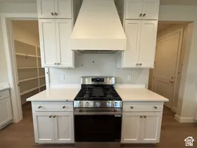 Kitchen featuring white shaker-style cabinetry, a custom vent hood, stainless steel gas range, solid-surface countertops, and an integrated walk-in pantry with built-in shelving