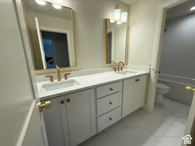 Dual-vanity bathroom featuring white cabinetry, a solid-surface countertop with integrated sinks, brass-toned hardware, rectangular gold-framed mirrors, and floor tile with a natural-stone pattern