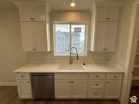 White shaker-style cabinetry with crown molding, light-toned solid surface countertops, stainless steel integrated dishwasher, undermount farmhouse sink with high-arc gooseneck faucet, and wood-finish flooring
