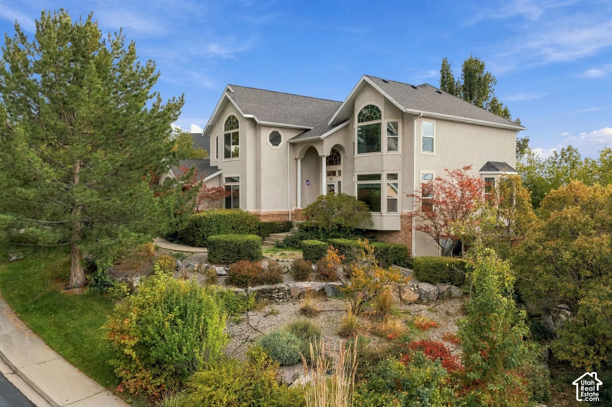 Two-story residence featuring a neutral-toned stucco exterior, multi-gabled roofline, arched window details, stone retaining wall hardscaping, and tiered landscaping