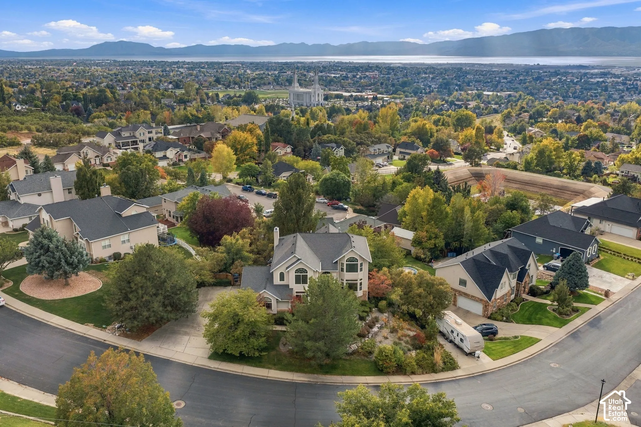 Aerial perspective of a hillside residential neighborhood featuring expansive mountain range backdrops, mature trees, multi-level residential architecture, paved cul-de-sac streets, and panoramic valley terrain