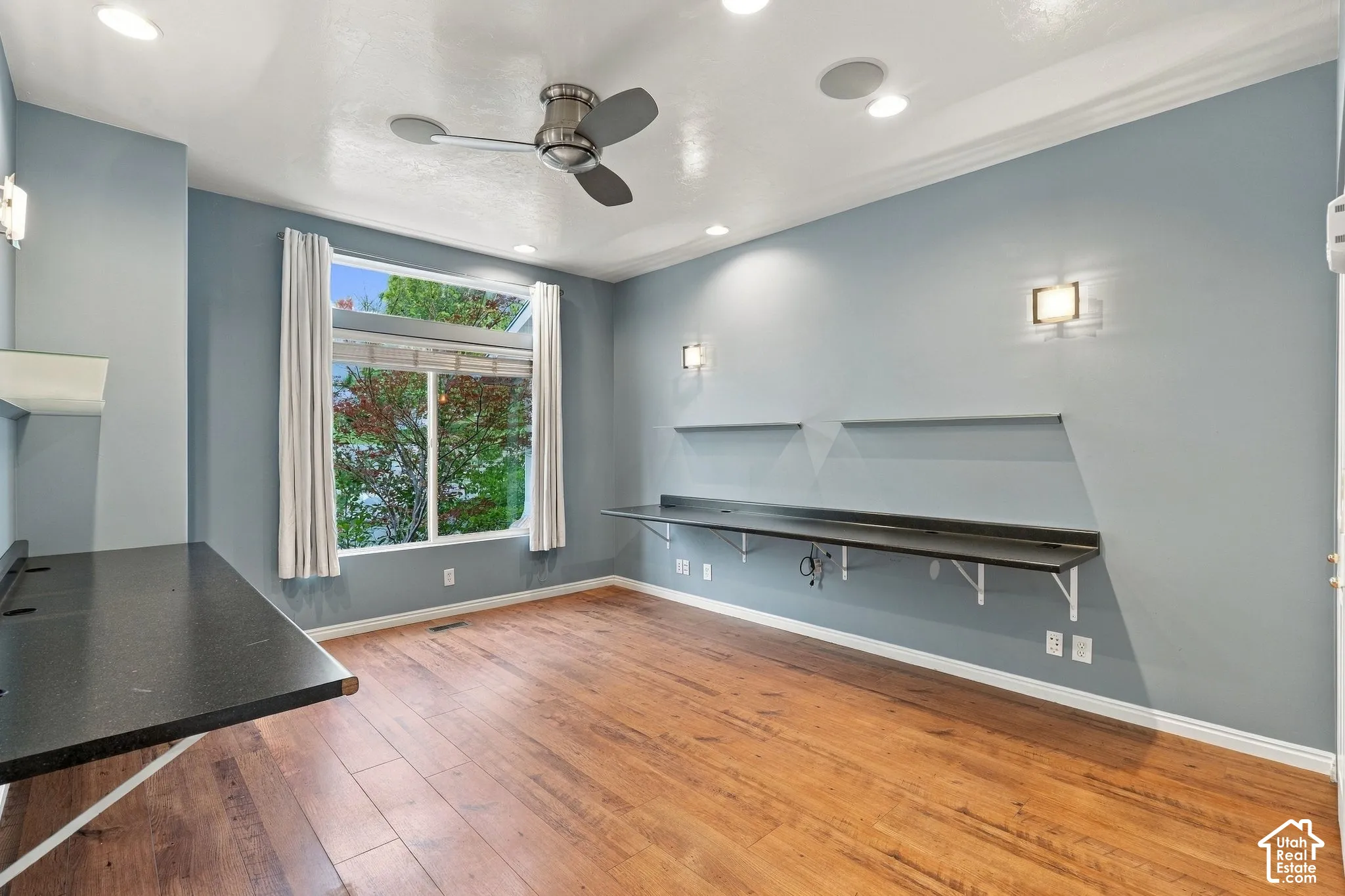 Room featuring wood-finish flooring, recessed lighting, a ceiling fan, dual wall-mounted desks with integrated shelving, and large windows
