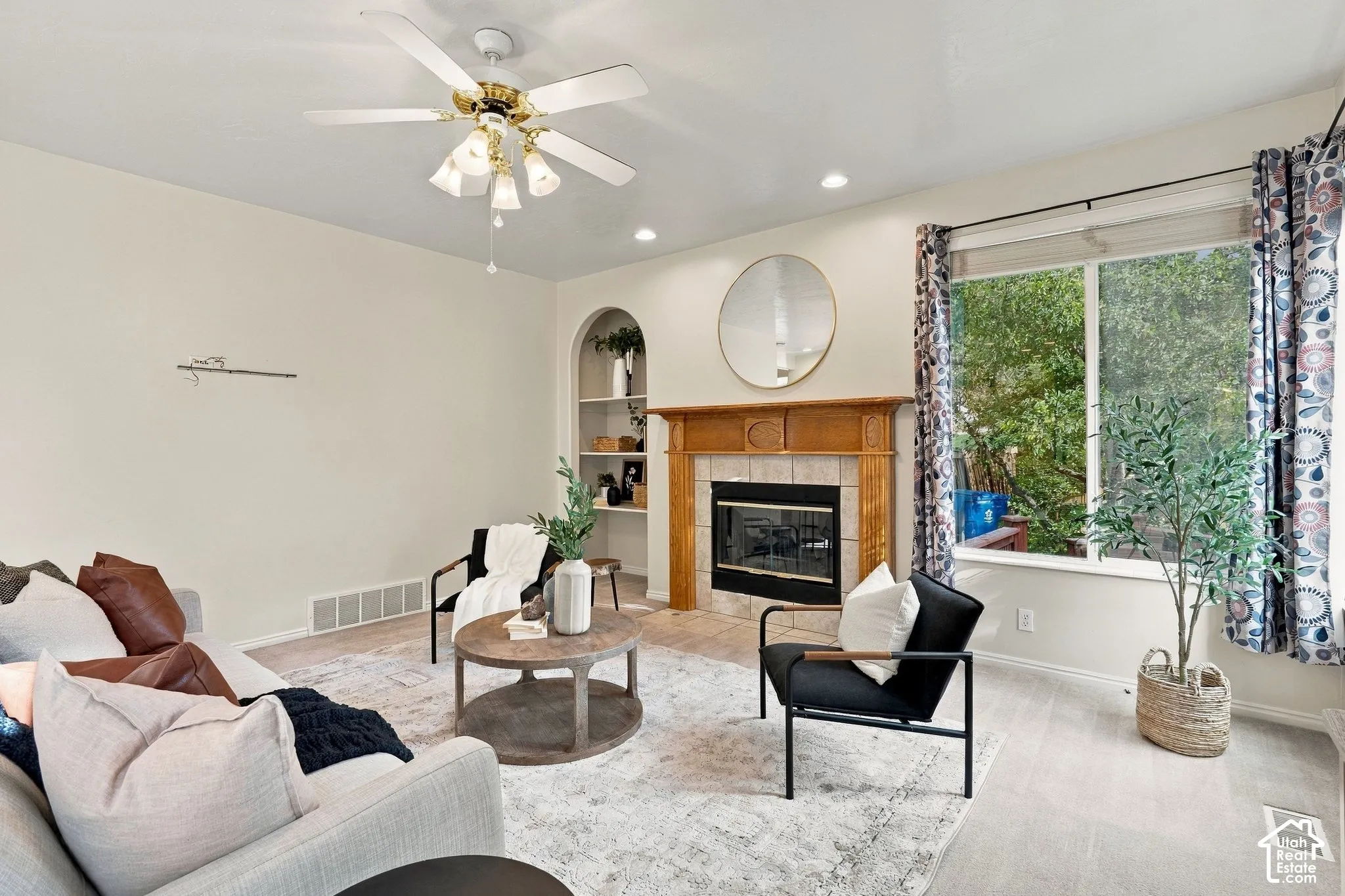 Living area featuring a fireplace with a wood mantel, tile surround, recessed arched shelving, a ceiling fan with integrated lighting, and large windows