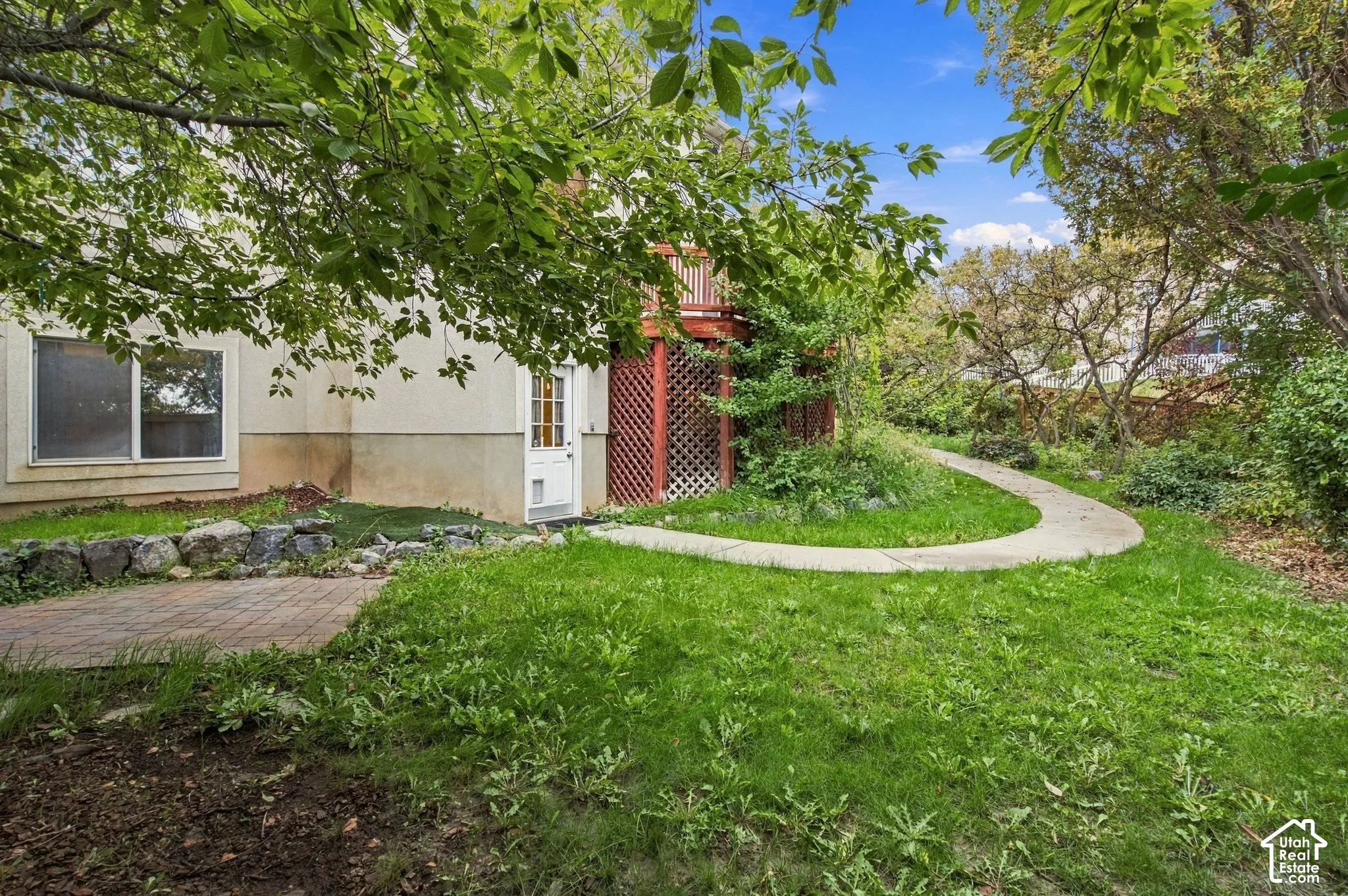 Rear exterior featuring a brick paver patio, stone retaining wall, elevated wood deck with lattice screening, and a curved concrete walkway