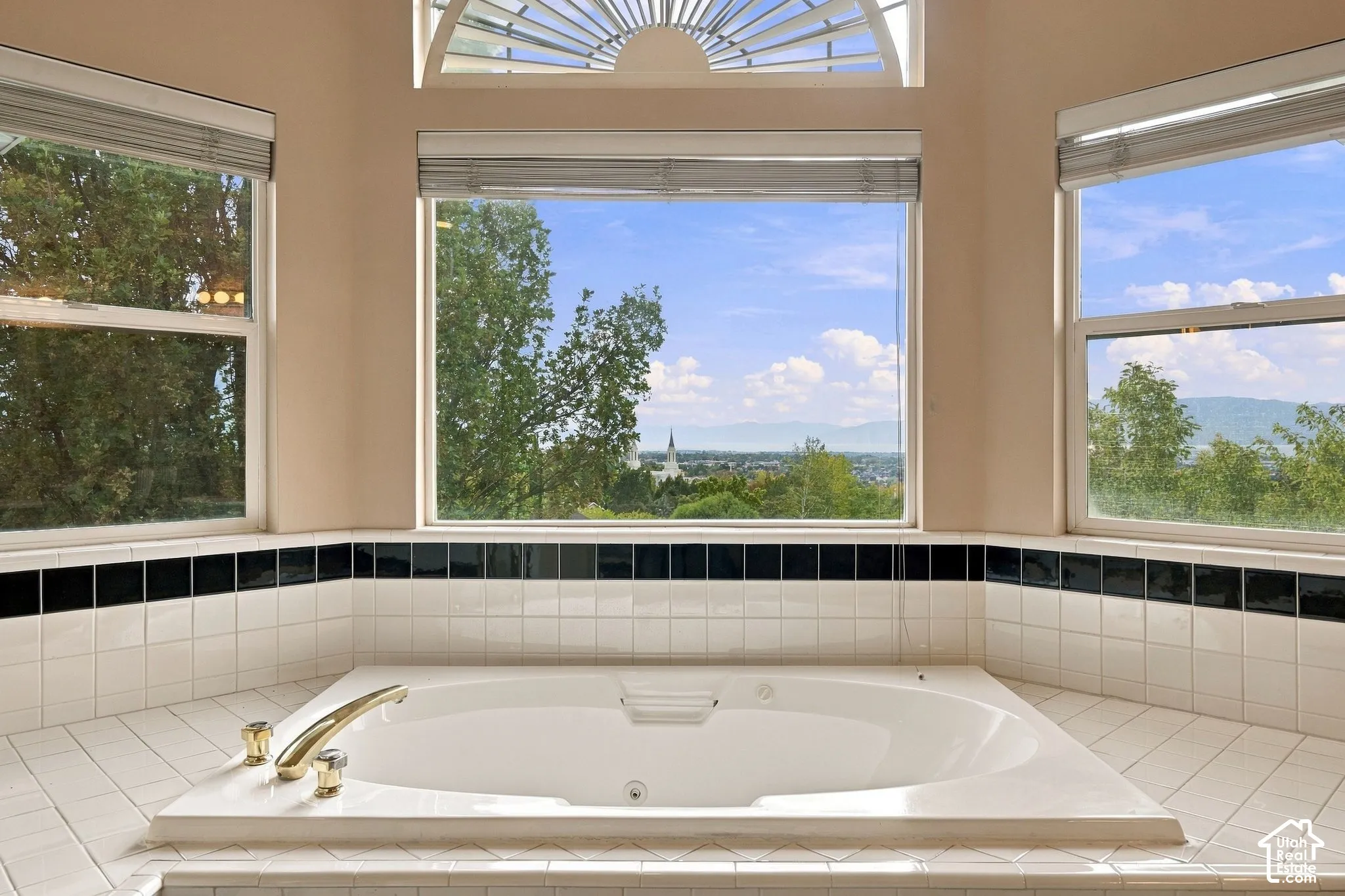 Built-in soaking tub with gold-tone fixtures, tile surround featuring black accent detailing, and expansive windows with arched transom overhead