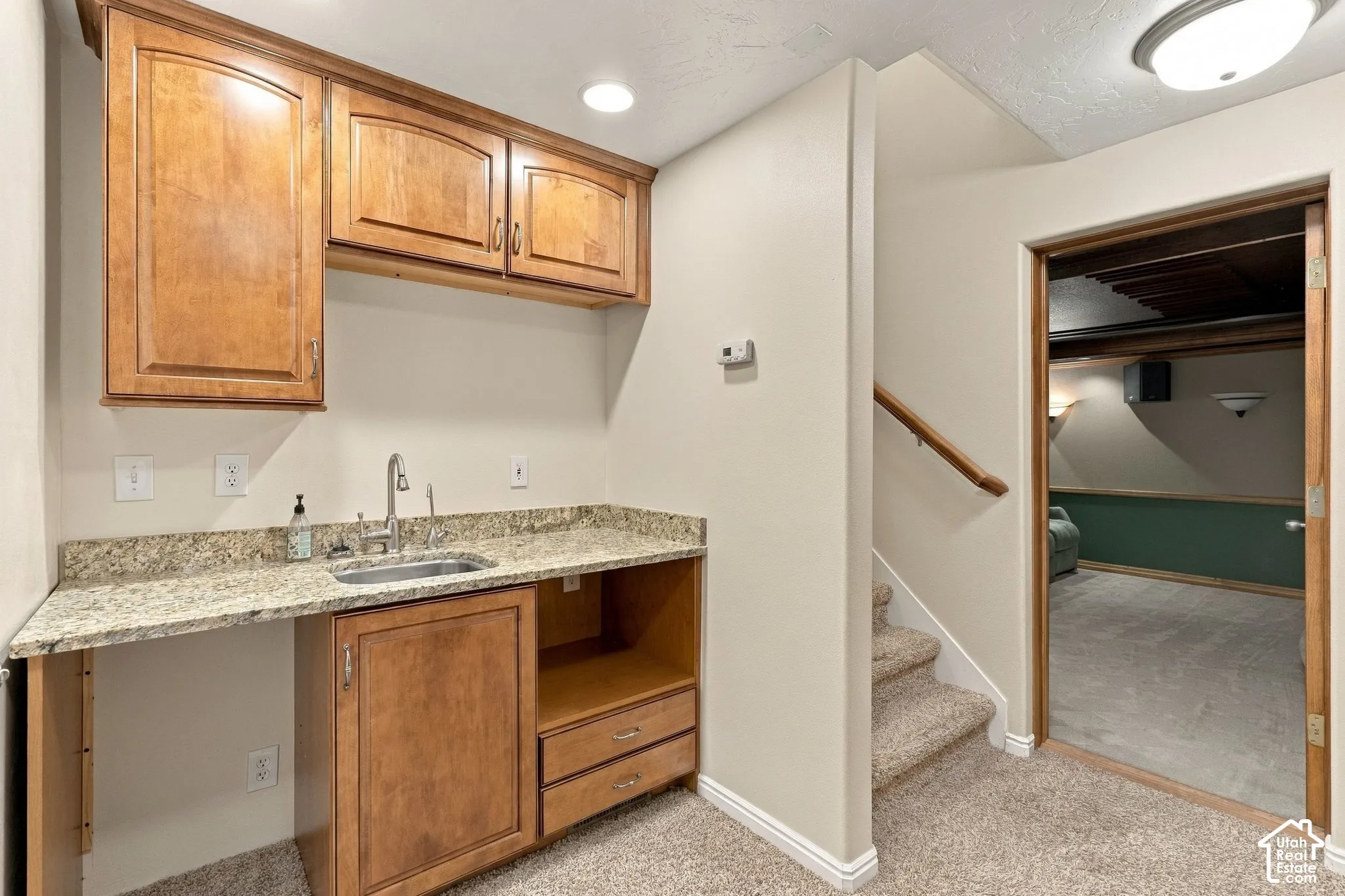 Wet bar featuring wood cabinetry, stone-surface countertop, stainless steel sink with gooseneck faucet, and carpeted stairway