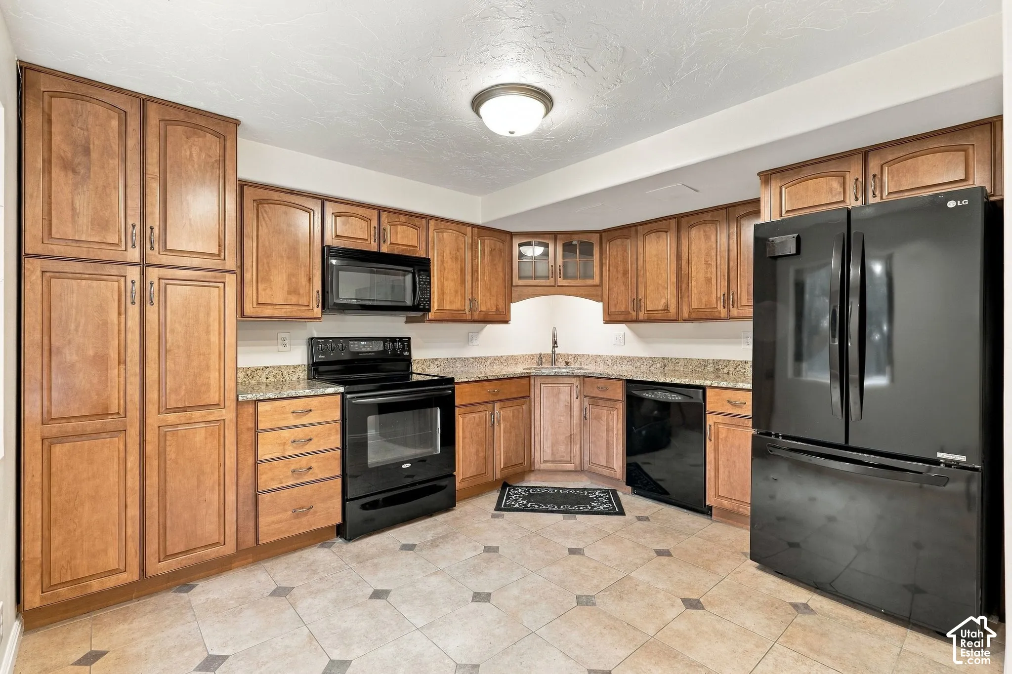 Kitchen featuring medium-toned wood cabinetry, stone countertops, black appliances, glass-front upper cabinet accents, and tile flooring