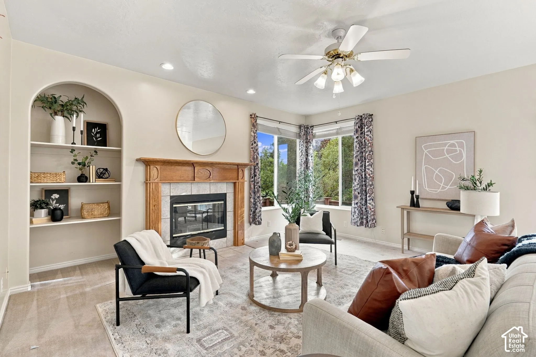 Living area featuring a fireplace with a wood mantel, tile surround, recessed arched shelving, large windows, and a ceiling fan with integrated lighting