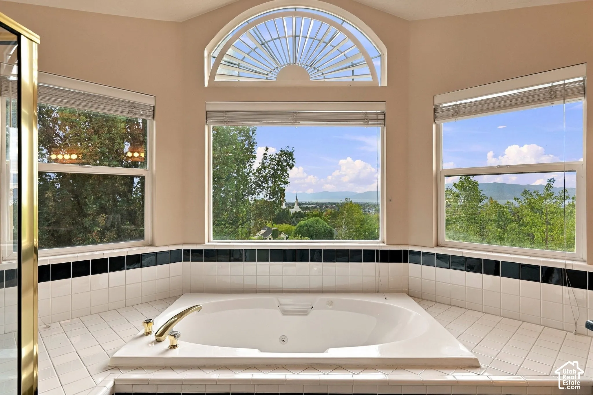 Jetted soaking tub with brass-finish fixtures, surrounded by white and black tile work, set within a bay window configuration featuring an arched transom and mountain scenery