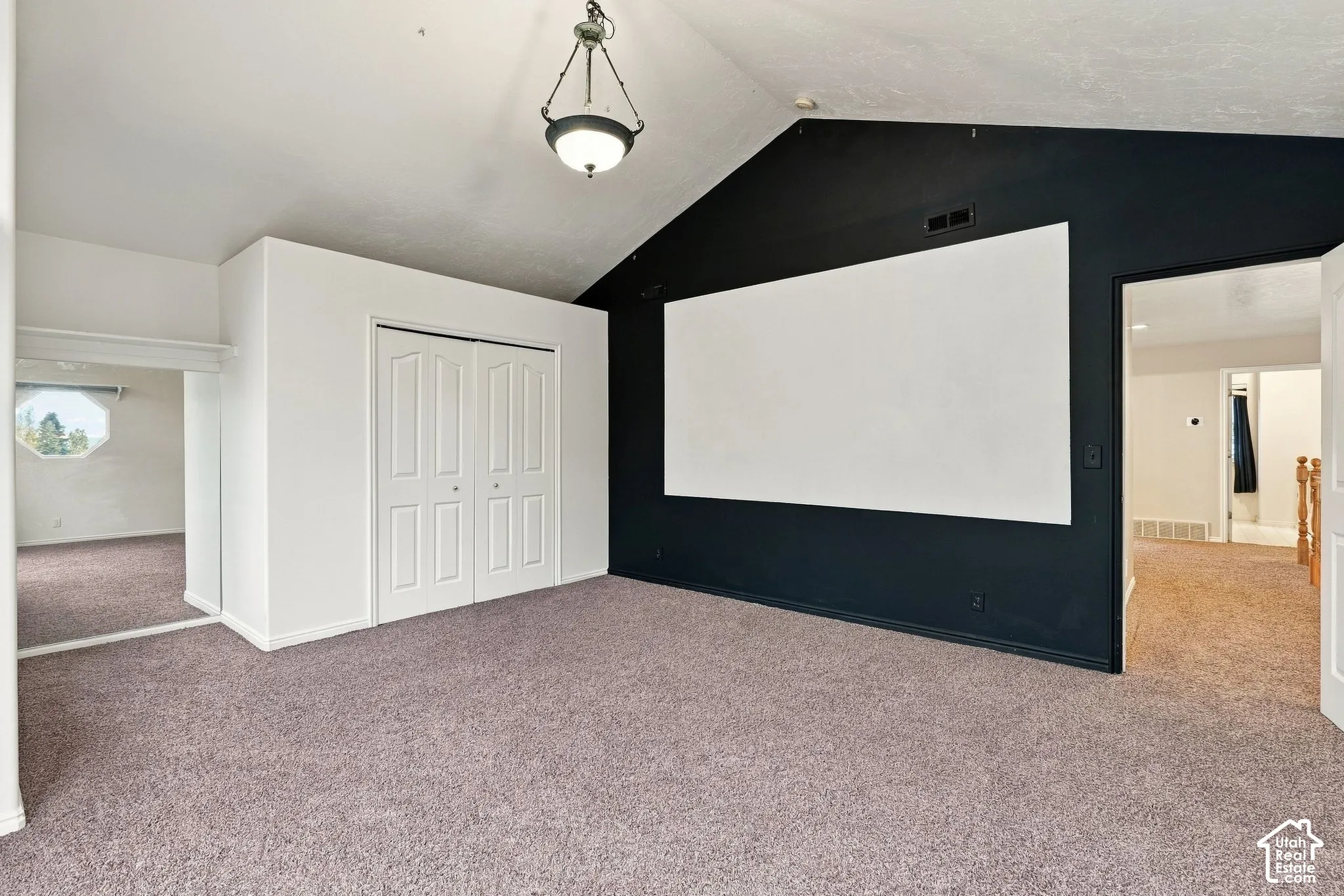 Vaulted ceilings, wall-mounted projection screen, neutral carpeting, dual-panel closet doors, and a decorative pendant light fixture