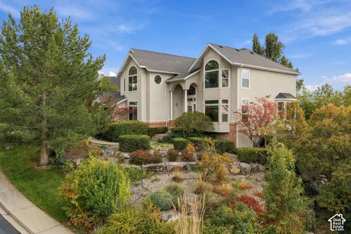 Two-story residence featuring a neutral-toned stucco exterior, multi-gabled roofline, arched window details, stone retaining wall hardscaping, and tiered landscaping