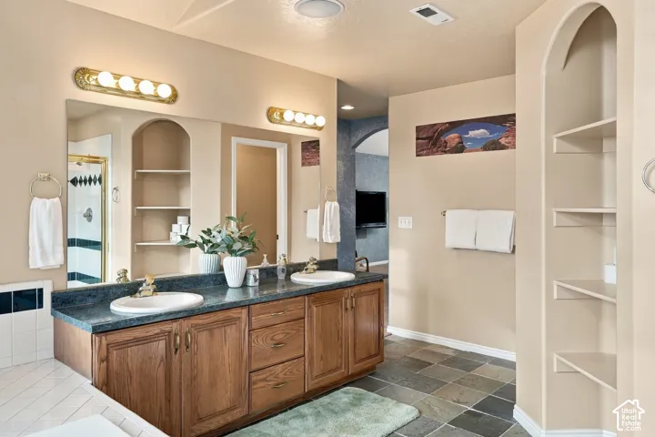 Dual-sink vanity with stone-finish countertop and wood cabinetry, wall-mounted light fixtures, large vanity mirrors, and built-in arched wall shelving