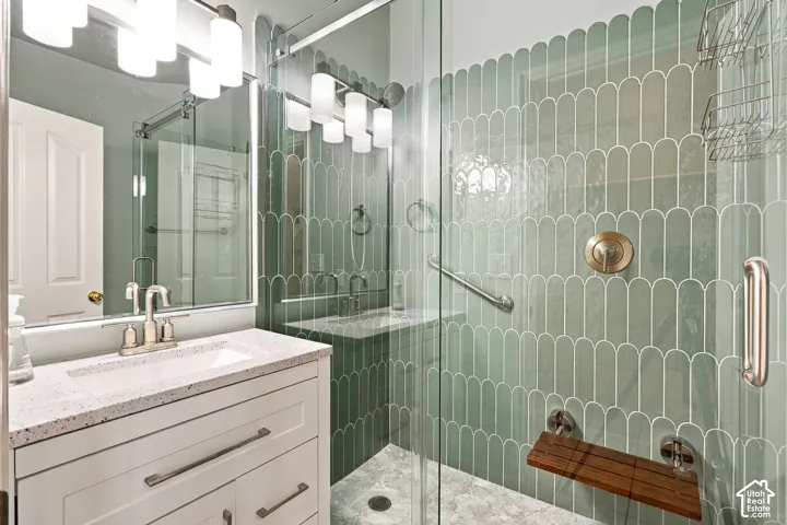 White vanity with speckled stone countertop, integrated rectangular sink, glass-enclosed shower with decorative scalloped tile, metallic hardware, and built-in folding seat