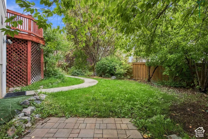 Rear yard featuring a raised wood deck with lattice skirting, a paved brick patio, a concrete path, and mature tree landscaping enclosed by a wooden fence