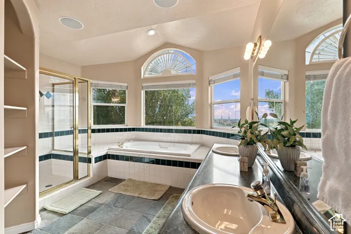 Bathroom featuring a deep soaking tub, separate glass-enclosed shower, dual-sink vanity with dark stone-surface countertops, tile flooring, and arched windows with fanlight accents
