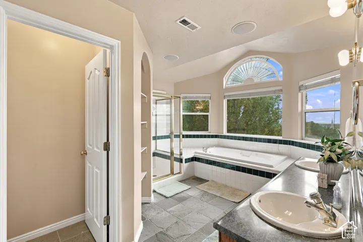 Dual-sink vanity with stone-finish countertop, soaking tub, glass-enclosed shower, vaulted ceiling, and arched window
