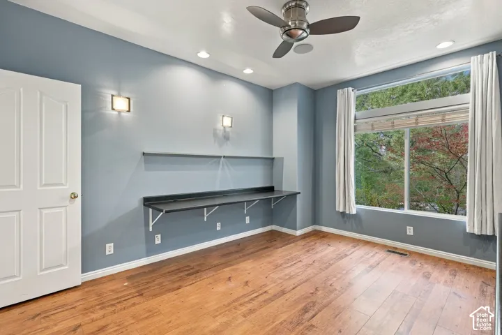 Wood-finish flooring, built-in desk workstation, wall-mounted light fixtures, multi-pane window, and brushed-nickel ceiling fan