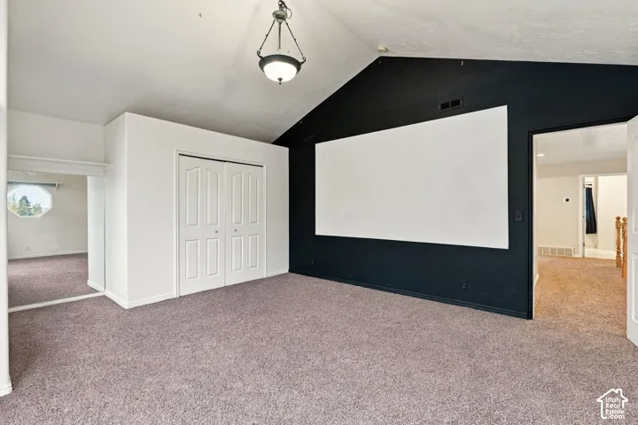 Vaulted ceilings, wall-mounted projection screen, neutral carpeting, dual-panel closet doors, and a decorative pendant light fixture