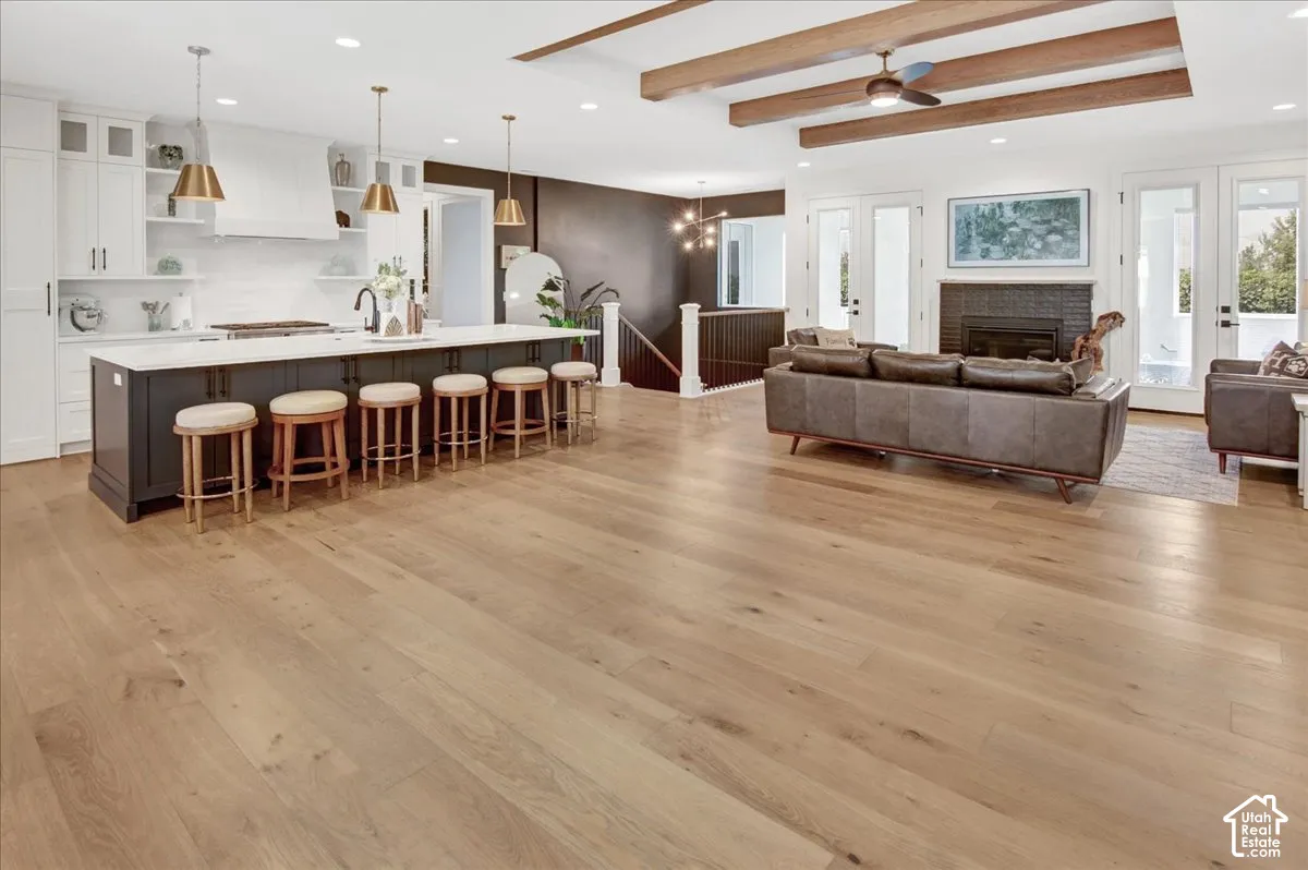 Open-concept living space with wood-finish flooring, a kitchen island with seating, exposed ceiling beams, a built-in fireplace, and double French doors