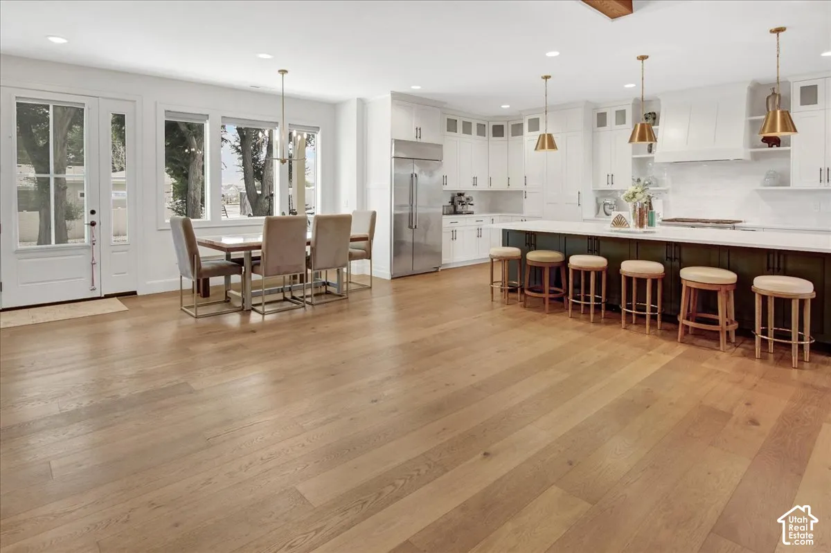Expansive open-concept space featuring wood-finish flooring, a kitchen island with seating, white shaker cabinetry, stainless steel appliances, and a dining area with large windows
