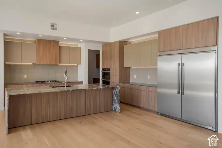 Kitchen featuring wood-finish cabinetry, a large waterfall-edge island, stone countertops, stainless steel appliances, and light wood-finish flooring