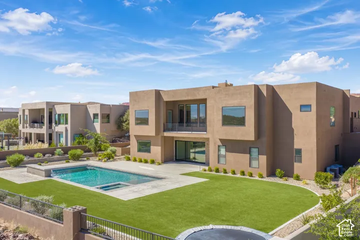 Modern stucco exterior featuring a two-story design, swimming pool with built-in spa, paved patio, synthetic lawn, and a second-story balcony with glass railing