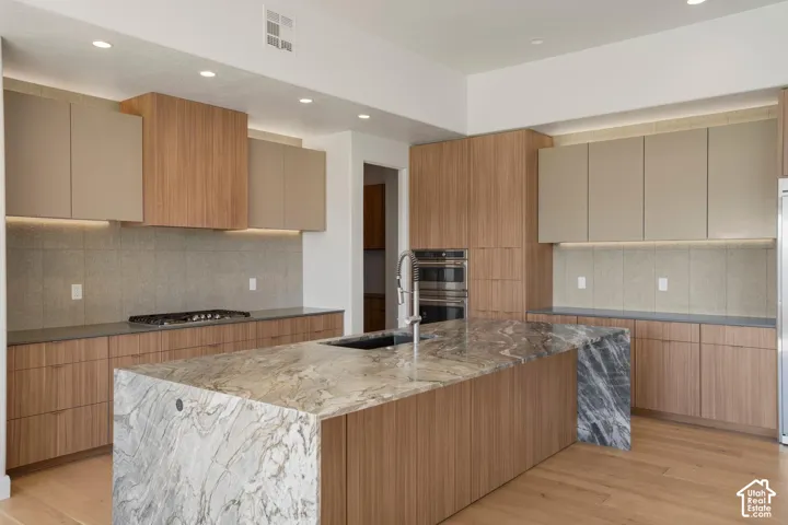 Kitchen featuring a waterfall-edge stone island, wood-finish cabinetry, built-in double wall ovens, a gas cooktop, and under-cabinet lighting