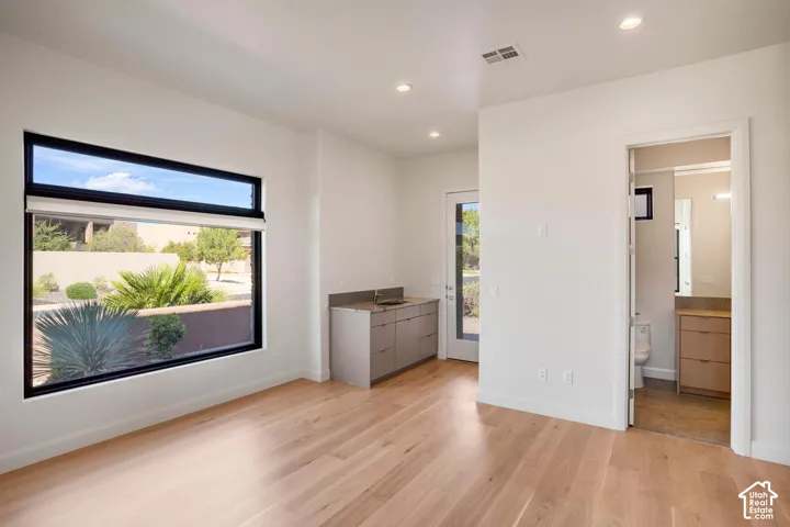 Wood-finish flooring, large rectangular window with dark trim, built-in kitchenette cabinet with integrated sink and stone countertop, and recessed ceiling lighting