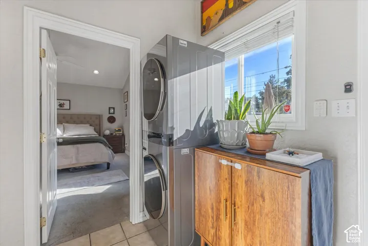 Utility area featuring a stacked stainless steel laundry unit, neutral tile flooring, and a window with horizontal blinds
