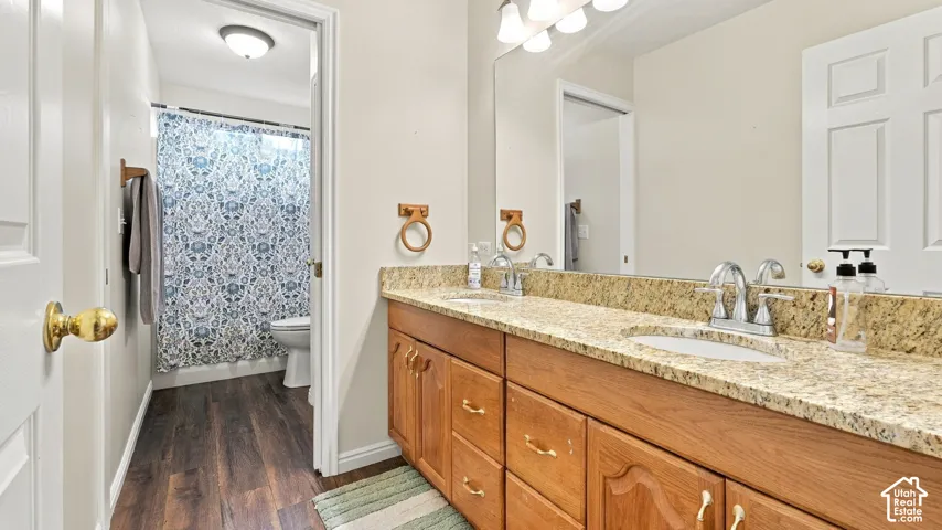 Double-vanity cabinetry with wood-finish doors, stone-composite countertop, dual undermount sinks, wide-format wall mirror, and wood-finish flooring