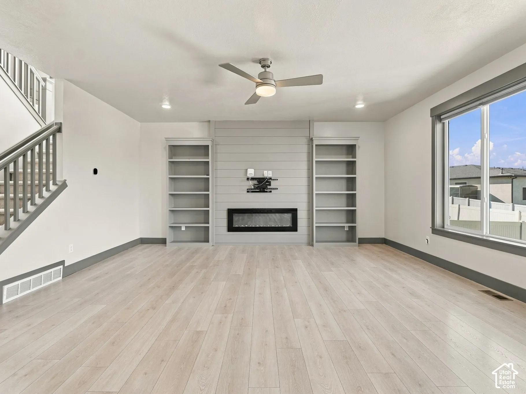 Living area featuring wood-finish flooring, a wall-mounted electric fireplace, built-in shelving, a modern ceiling fan, and a staircase with a gray balustrade