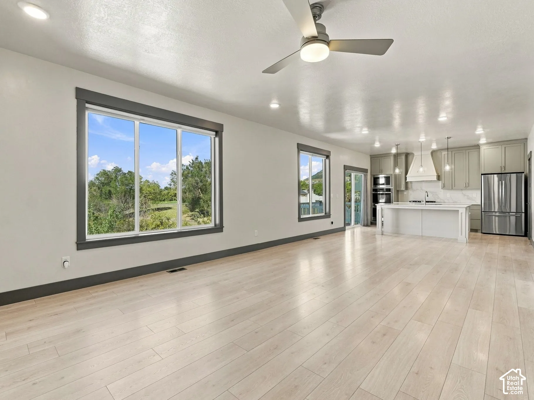 Open-concept interior featuring light wood-finish flooring, recessed lighting, a modern ceiling fan, large windows with dark trim, and a kitchen equipped with a central island and stainless-steel appliances