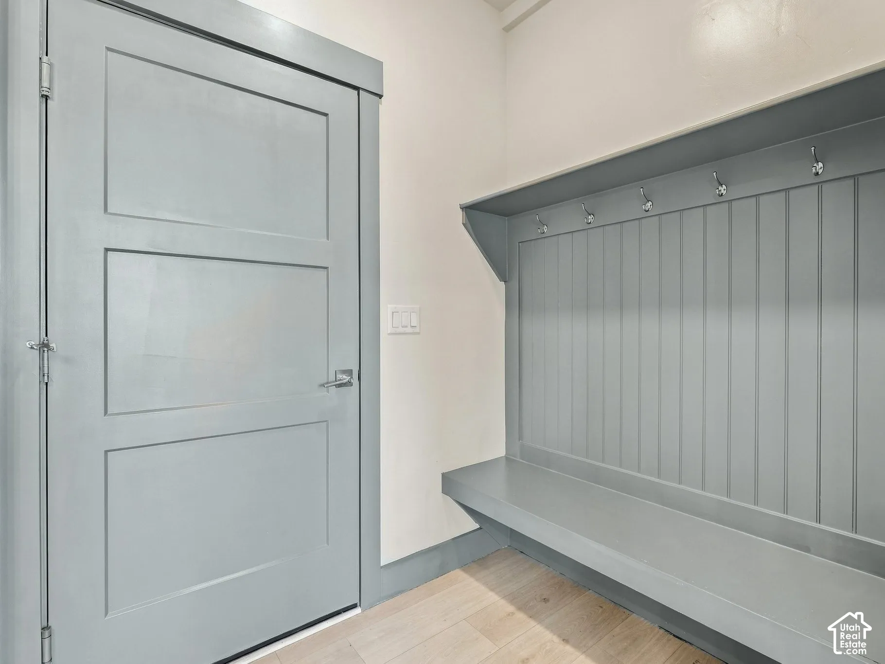 Built-in mudroom bench with beadboard wainscoting, overhead shelf, metal garment hooks, and three-panel door finished in coordinating gray paint, complemented by light wood-finish flooring
