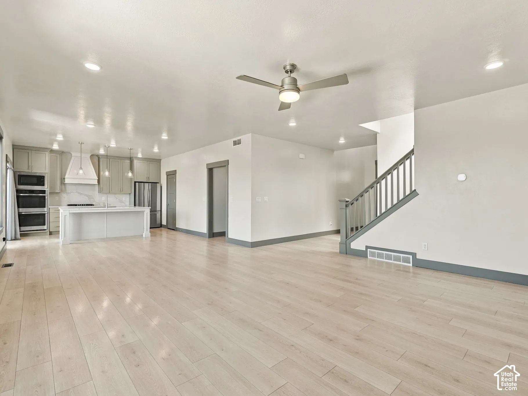Open-concept area featuring light-toned wood-finish flooring, a kitchen with a built-in double oven and center island, recessed ceiling lighting, and a staircase with a wooden handrail