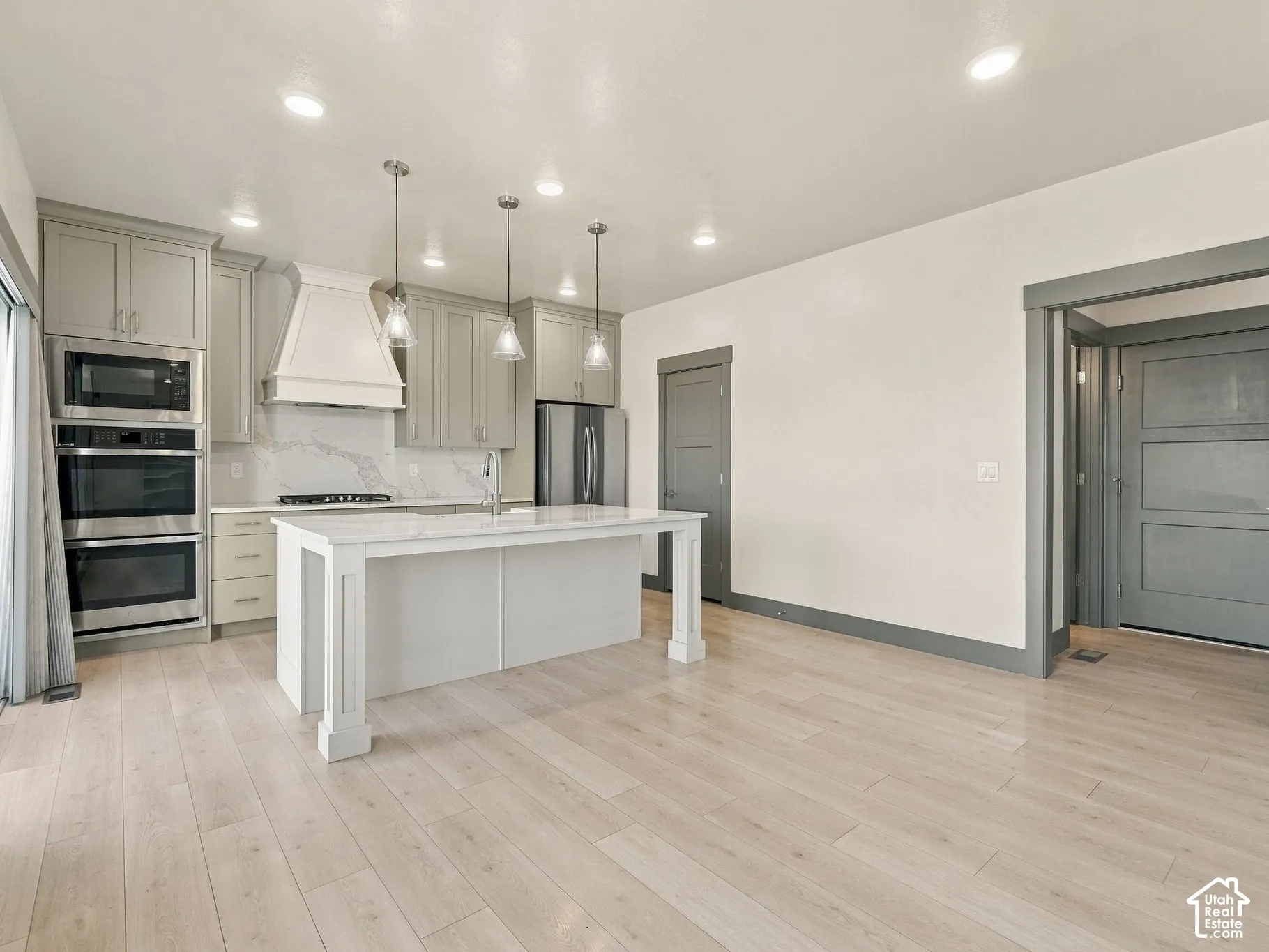 Kitchen featuring light-toned wood-finish flooring, a central island with a quartz countertop, custom cabinetry, stainless steel double wall ovens, and pendant lighting