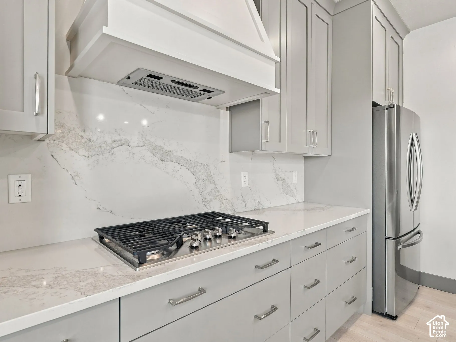 Kitchen featuring gray shaker-style cabinetry, a stone-patterned backsplash, quartz countertops, a stainless steel gas cooktop with a custom range hood, and a stainless steel refrigerator