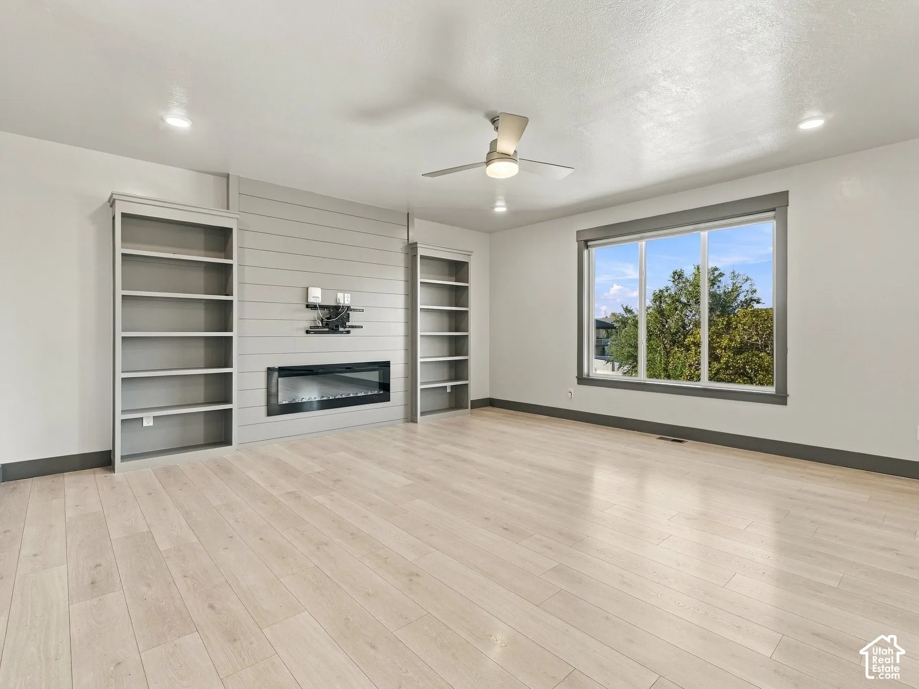 Light-toned wood-finish flooring, linear electric fireplace set in a shiplap feature wall, built-in shelving, large picture window, and ceiling-mounted fan